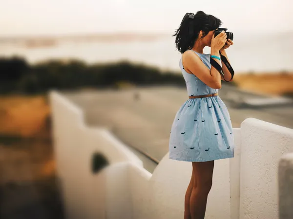 A woman photographer wearing a blue sleeveless dress takes a photograph for a free stock photography website.