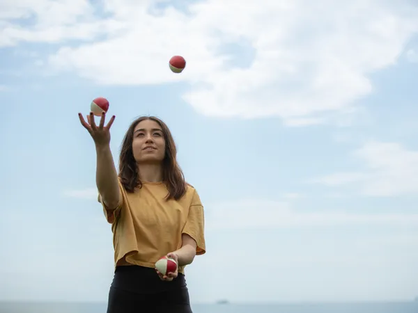 A woman standing trying to juggle, she’s holding two balls and is looking at the third ball in the air. 