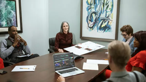 A woman sitting on a black chair holding a board in a conference room with a group of people.